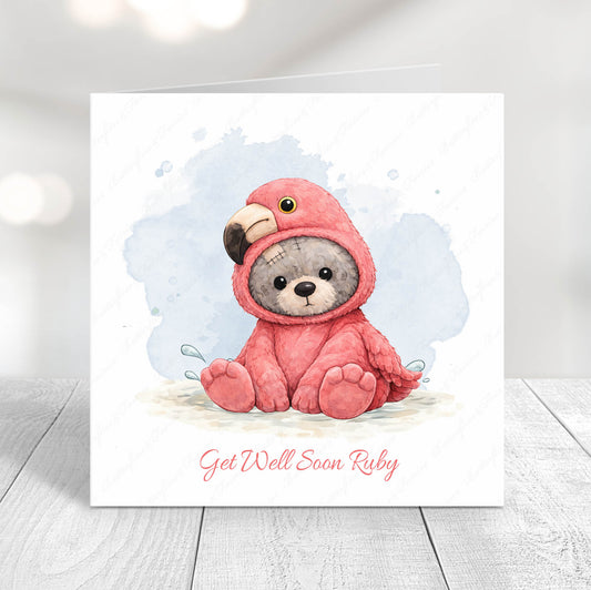 Personalised get well soon card featuring a cute teddy bear wearing a pink flamingo costume, sitting on a soft blue watercolour background, with the message “Get Well Soon Name” underneath.